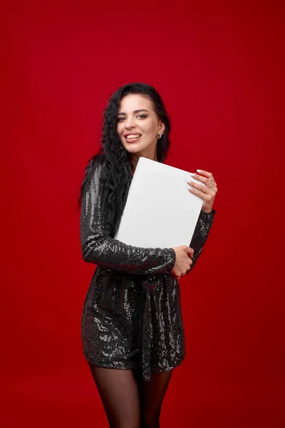 An elegant girl in a black short dress on a red background hugs an empty white box and looks at the camera. Advertising your product in the arms of a girl. Discount advertising.