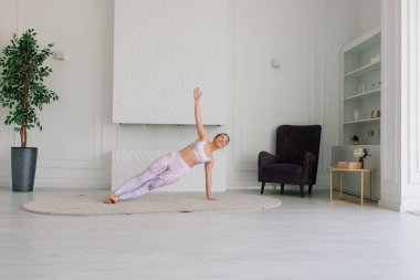 Genç, çekici, gülümseyen bir kadın yoga yapıyor, spor kıyafetleri giyiyor, sutyen giyiyor, evde eğitim görüyor.