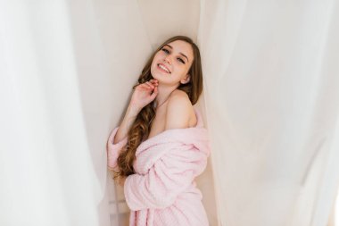 Evin içinde, büyük pencerelerin yanında beyaz bir bornozla poz veren güzel, harika bir genç kadın resmi..