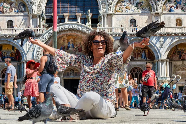 Venedik, İtalya - 25 Ağustos 2021 'de güvercinler gülümseyen bir Brezilyalı kadının ellerine oturur ve ekmek yerler. San Marco Meydanı 'nda turistler arasında meşhur bir tören.