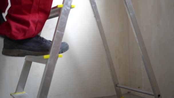 Malíř v práci s válečkem, kýblem a žebříkem. Šedovlasý zralý stavební dělník v červené kombinéze vyleze po žebříku a začne malovat strop bílou barvou — Stock video