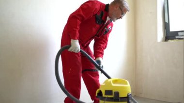 Turuncu tulumlu ve gözlüklü işçi inşaat işlerini süpürüyor ve odada elektrikli süpürgeyle toz tutuyor. Statik çekim