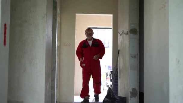 Olgun gri sakallı, kırmızı tulumlu inşaat işçisi yeni bir binaya giriyor. Emlak ve inşaat, iç işleri konsepti. — Stok video