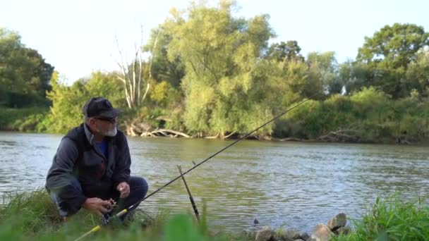 Elderly gray-bearded fisherman sets up spinning, fishing line and tackle on a lake in the countryside. Weekend activity, out-of-town fishing — Stock Video