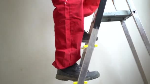 Hombre pintor en el trabajo con un rodillo, cubo y escalera. trabajador maduro de la construcción de pelo gris en mono rojo sube una escalera y comienza a pintar el techo con pintura blanca — Vídeos de Stock