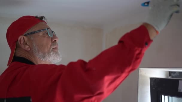 Porträt eines graubärtigen Bauarbeiters in einem roten Overall, der die Decke bemalt, während er auf einer Leiter steht — Stockvideo