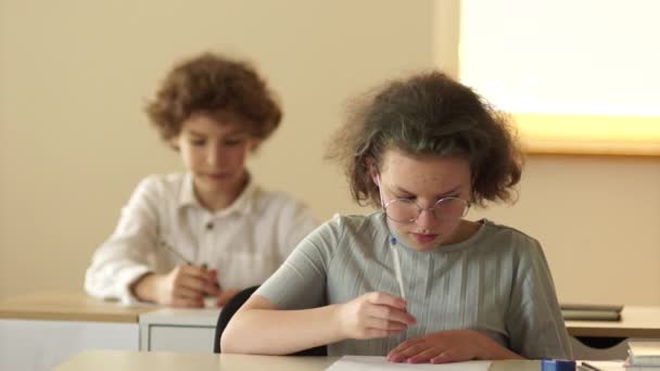 Dospívající dívka s brýlemi se vesele směje ve škole během lekce. Ta školačka má za stolem vějíř. Šťastné školáky, zpátky do školy — Stock video