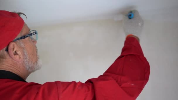 La capa de pintura madura pinta el techo de blanco. Un hombre con un mono rojo. Concepto de obras de renovación y acabado, inmobiliaria e hipotecaria — Vídeos de Stock