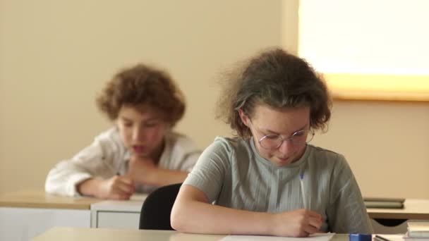 Een gekruld schoolmeisje met een bril schrijft een test en gluurt naar een spiekbriefje. Het meisje lacht terwijl ze aan haar bureau zit in de klas. Leraren handen pick-up vellen papier — Stockvideo