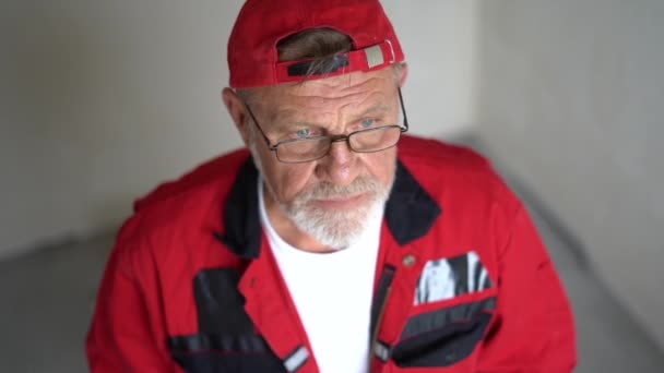 Porträt eines älteren Ingenieurs in rotem Overall und Mütze mit grauem Bart und Brille, der in die Kamera blickt und lächelt — Stockvideo