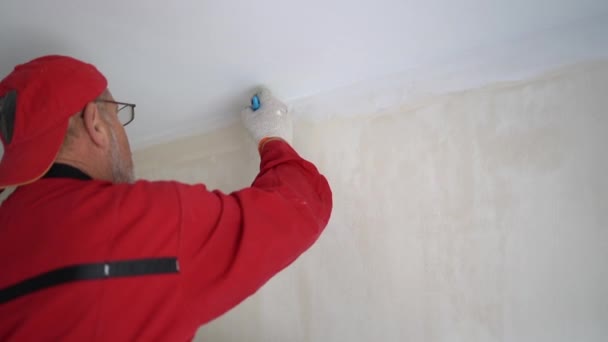 Renovación y decoración interior de una casa nueva. Un trabajador en mono rojo pinta el techo con pintura blanca — Vídeo de stock