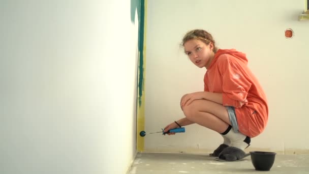 Menina pintando a parede e olhando para a câmera sorrindo. Rapariga pinta a parede com uma almofada enquanto se agacha. Uma adolescente com cabelo encaracolado vermelho faz reparos em seu quarto — Vídeo de Stock