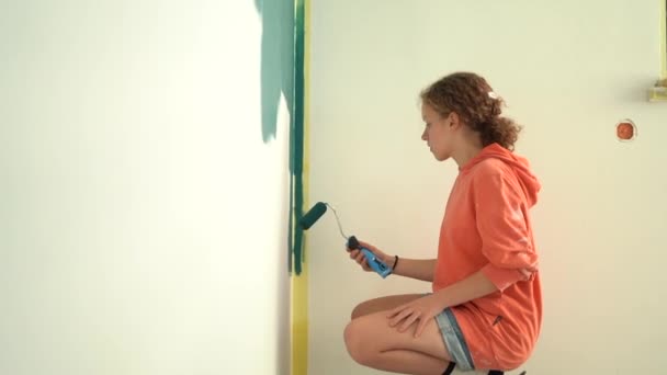 Koncept rekonstrukce a kreativity. Dívka se mu posadí na paty a přikrčí se u stěny s malířským nářadím. Teenage dívka s červenými kudrnatými vlasy maluje zeď modrá s válečkem ve svém vlastním pokoji — Stock video