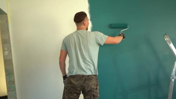 Ένας ενήλικας κάνει επισκευές μόνος του στο σπίτι. Ο τύπος βάφει τον τοίχο με ένα μεγάλο ρολό, επισκευαστικό σχέδιο. Πίσω όψη — Αρχείο Βίντεο