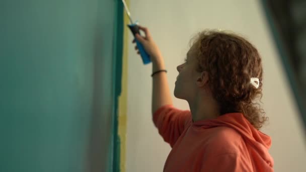 戴眼镜的女孩把一个滚筒浸在油漆中，然后爬上梯子，在她家粉刷墙壁。修理的概念。公寓里的年轻女人画墙壁 — 图库视频影像