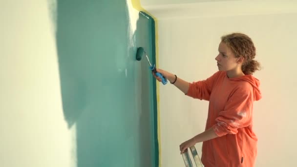 Вид сбоку молодой женщины, стоящей на лестнице, раскрашивает стену кисточкой синего цвета. Концепция реконструкции — стоковое видео