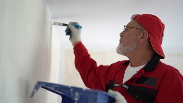 退職者は白い塗料で壁をペイントします。赤い帽子を被った灰色の髭を生やした男が新しい家で修理をする — ストック動画