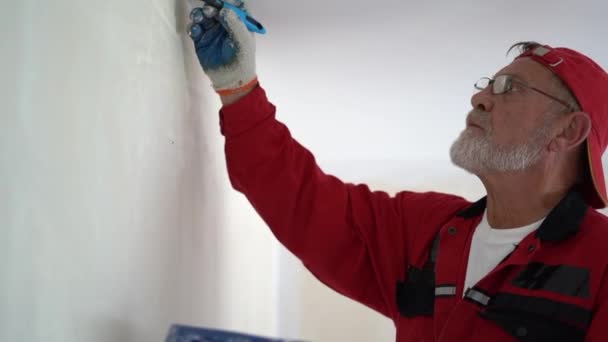 Renovering och fastigheter koncept, njuta av arbetet. Gammal gråskäggig man i röd mössa och glasögon målar väggen med en vit pensel i sin nya lägenhet — Stockvideo