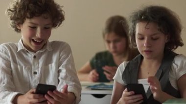 Ciddi kıvırcık saçlı okul çocukları, bir oğlan ve bir kız, okuldaki çocuklar ders sırasında akıllı telefonlarının ekranlarına bakarlar. Oğlan kıza telefonunu gösteriyor. Çocuklar ve aletler