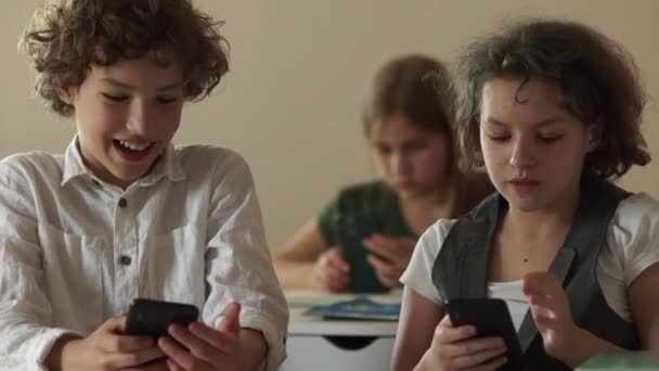 Los escolares serios de pelo rizado, un niño y una niña, los niños en la escuela miran las pantallas de sus teléfonos inteligentes durante la lección. El chico le muestra su teléfono a la chica. Niños y aparatos — Vídeo de stock