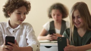 Sınıf arkadaşları, bir erkek ve bir kız, okul masasında oturuyorlar ve akıllı telefonlarının ekranlarına bakıyorlar. Okula dönün, çocuklar ve aletler.