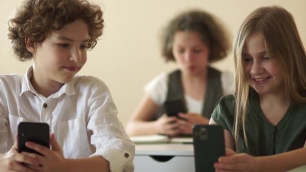 Klasgenoten, een jongen en een meisje, zitten aan een schoolbureau en kijken naar de schermen van hun smartphones. Terug naar school, kinderen en gadgets — Stockvideo
