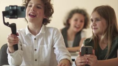 Komik okul çocuğu portresi. Blog yazarı, kıvırcık saçlı, genç bir çocuk. Bir okul çocuğu bir okul sınıfından bir dereyi kaydediyor. Video günlüğü ve hikaye kavramı