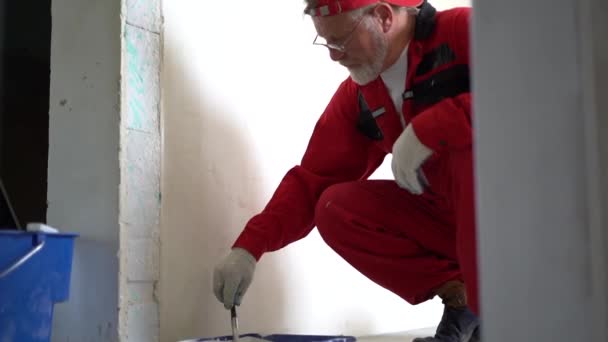 Oudere bebaarde bouwvakker in rode overall hurkend met een roller met witte verf op de muur — Stockvideo