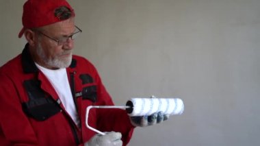 Duvarları boyamak için yeni silindirli kırmızı tulumlu ve gözlüklü yaşlı gri saçlı bir inşaat işçisinin portresi. Yenileme ve emlak konsepti