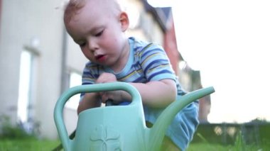 Şirin sarışın bebek hortumdan plastik bir sulama kutusuna su döküyor. Yaz tatili. Mutlu çocuk suyla oynuyor.