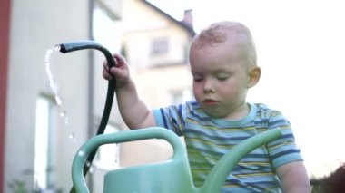 Küçük çocuk bir hortum ve büyük bir plastik sulama kutusuyla dışarıda oynuyor. Sarışın çocuğun yazın köyde bir hayranı varmış.