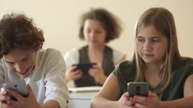 Sınıf arkadaşları, bir erkek ve bir kız, okul masasında oturuyorlar ve akıllı telefonlarının ekranlarına bakıyorlar. Okula dönün, çocuklar ve aletler.