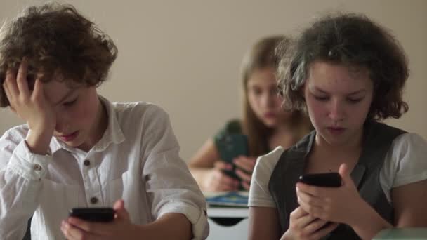 Çocuklar ve aletler. Okuldaki öğrenciler akıllı telefonlarıyla meşguller. Bir erkek ve bir kız bir masada oturup telefonlarından ekranlara bakıyorlar. — Stok video