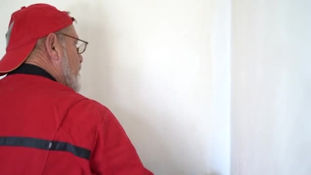 Portret dojrzałego pracownika w czerwonej czapce i kombinezonie malującym ścianę. Złota rączka pracuje w nowych nieruchomościach — Wideo stockowe