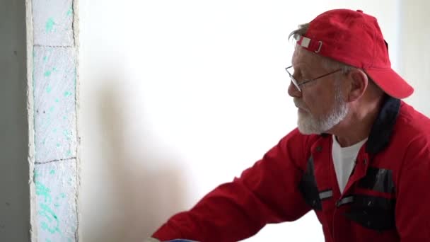 Close-up portret van charismatische grijze bebaarde volwassen bouwvakker met rode pet en overalls die de muur schilderen met witte verf — Stockvideo