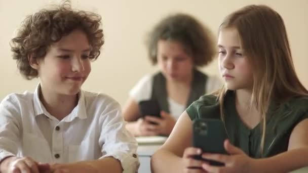 En vakker, langhåret tenåringsjente lager selfie med klassekameraten sin mens hun sitter ved pulten sin på skolen. Begrepet "barn og utstyr" – stockvideo