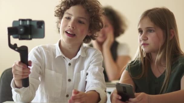 Mignon adolescent aux cheveux bouclés enregistre une vidéo pour son blog vidéo à son bureau dans la salle de classe. Un écolier tient dans ses mains un smartphone et un steadicam assis à côté d'un camarade de classe — Video