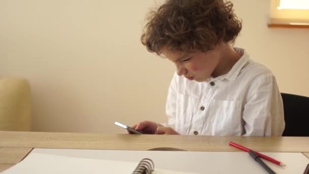 Schoolboy addicted to smartphones. Curly-haired teenage schoolboy in a white shirt is busy with his phone, texting friends during a break at school, sitting at his desk — Video Stock