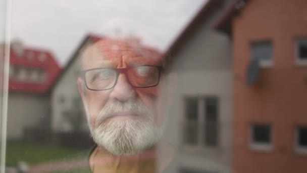 Retrato de um homem solitário de barba cinzenta na janela de sua casa. As casas são refletidas no vidro. Conceito de bloqueio e velhice — Vídeo de Stock