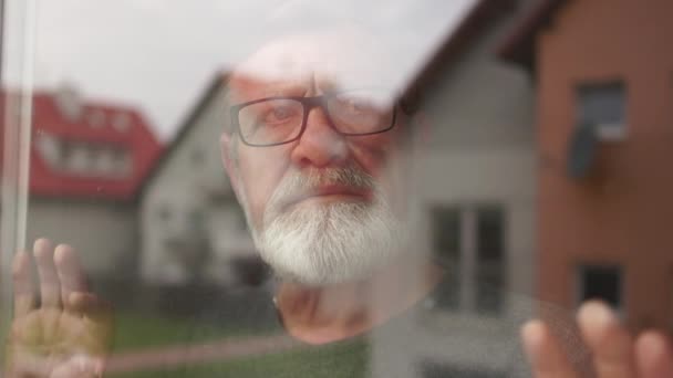 Одинокий человек печально смотрит в окно своего дома. Седобородатый пенсионер носит очки, кладет руки на стекло. Депрессия во время блокировки — стоковое видео