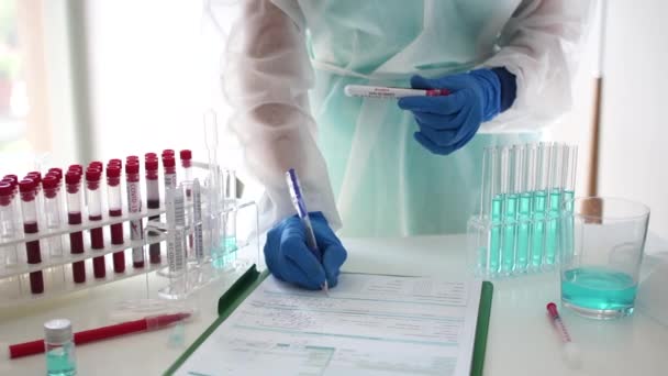 Невідомий лаборант у захисному костюмі та рукавичках фіксує результати тесту на коронавірус. Стійка з пробними трубами з зразками крові. Тест на ковід-19 — стокове відео