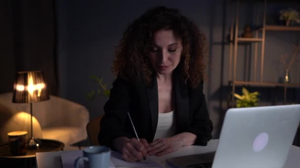 Una encantadora mujer de negocios que trabaja hasta tarde en la oficina. Mujer de pelo rizado en un traje de negocios termina el día de trabajo y cierra la computadora portátil — Vídeos de Stock