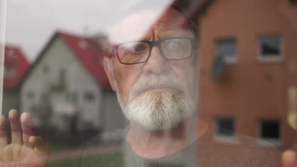 Lonely depressed gray-bearded elderly man with glasses stands by the window and looks into the distance. His wife comes up from behind to console — стоковое видео