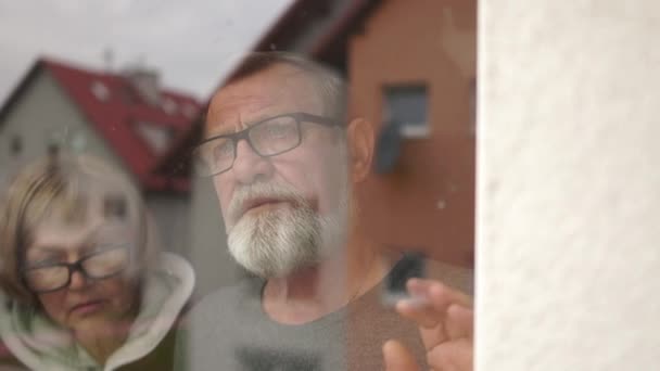 Sad couple of gray-haired pensioners, husband and wife, man and woman stand near the window during self-isolation covid-19 — Stockvideo