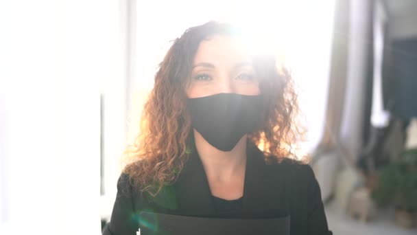 Солнечный портрет учителя с папкой в руках и в черной маске. Милая кудрявая девушка на фоне окна — стоковое видео