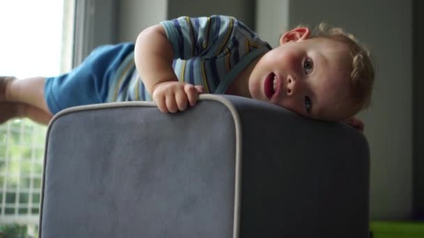 Portrait of a cute blond todler boy at home near the window. The child put his head on the ottoman and is resting. Happy childhood, a boy in kindergarten — Vídeo de Stock