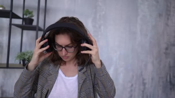 Young woman puts on headphones and prepares for a video call while sitting in the office near a laptop. Videoconference concept — Vídeos de Stock