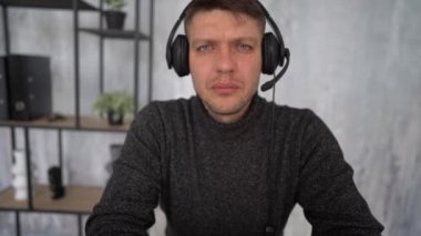 Video call. Portrait of a serious adult male businessman in a gray sweater with headphones on his head sitting at a table in the office