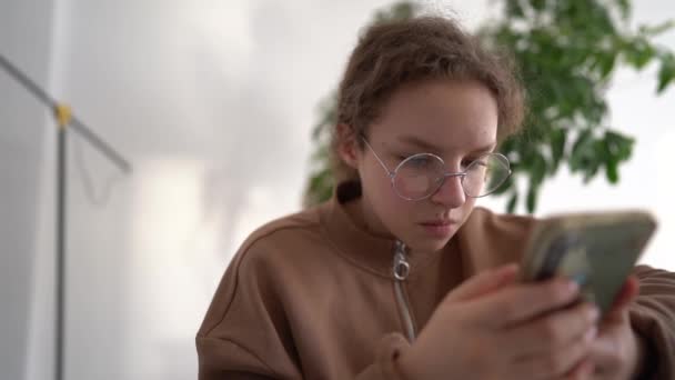 Έφηβη κοπέλα συμβουλεύεται τη φίλη της χρησιμοποιώντας μοντέρνο τηλέφωνο, ενώ κάθεται στο τραπέζι του γραφείου στο σαλόνι. Φοιτητής περιήγηση στον τρόπο ζωής πληροφορίες στο διαδίκτυο κατά τη διάρκεια της καραντίνας coronavirus — Αρχείο Βίντεο