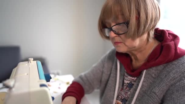 Pensjonistdame med briller syr tekstiler på en symaskin. Pensjonert kvinne og hennes hobby, bestemor jobber hjemme – stockvideo